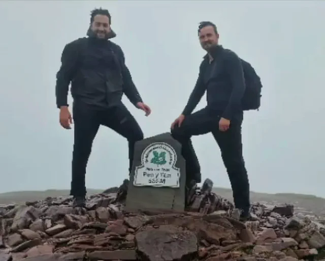 Jon and Josh at the summit marker on Pen y Fan, in mist, each with a foot on the stones.
