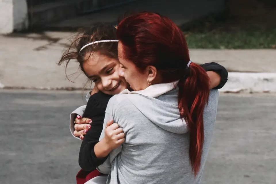 A mother in a grey hoodie scoops up her young daughter in the street, and the girl grins, her cheek pressed to her mother's