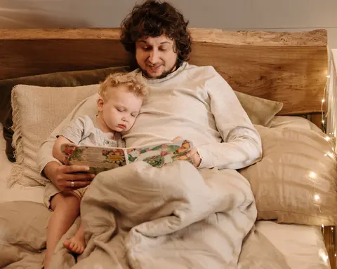 A father sits up in bed reading a picture book aloud, smiling, while his sleepy toddler drifts off against his chest at bedtime, early in the evening, in warm lamplight