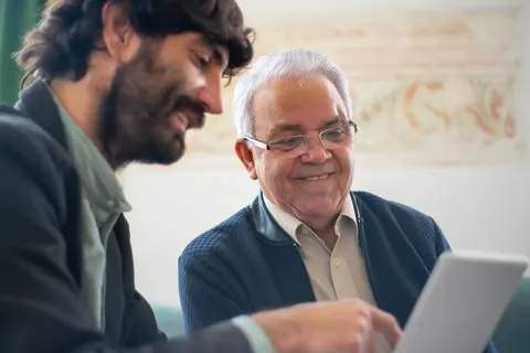 A bearded man and an older man in reading glasses sit side by side at a laptop, both smiling