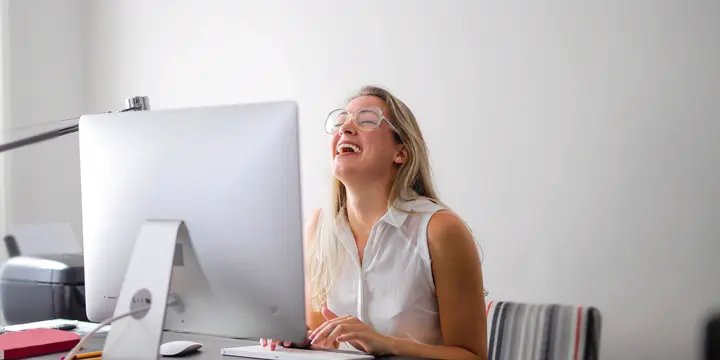 A woman laughs at her desk, her head thrown back, her screen in front of her, in bright window light