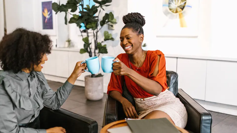 Two women laughing as they clink blue coffee mugs together in a bright lounge