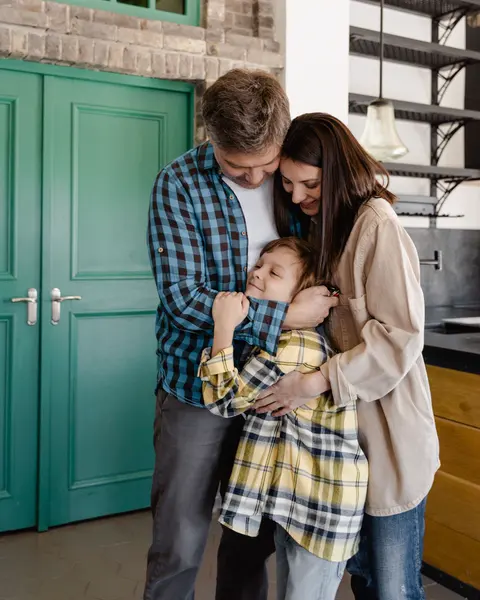 A parent home from work, hugging their partner and child in the kitchen