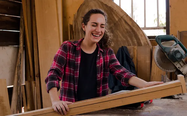 A young woman in a red check shirt laughs with her eyes shut at her workbench in a sunlit timber workshop, both hands on a long plank of wood