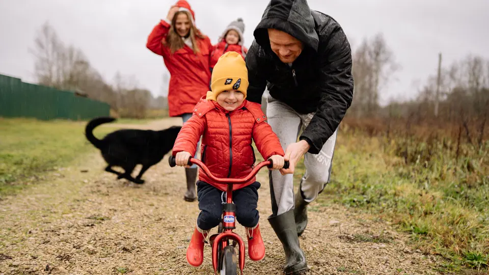 A parent in wellies helping a small child ride a bike down a country lane, the family and their dog just behind