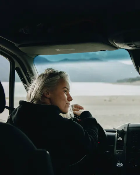 A woman sits in the driver's seat of her parked van at the end of the working day, arms on the wheel, looking out with a quiet half smile