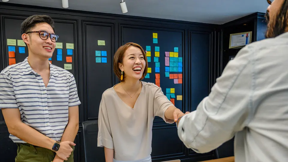 In a small studio with sticky notes all over a dark wall, a woman laughs as she shakes a new team member's hand on his first day, while a colleague in a striped shirt grins beside her