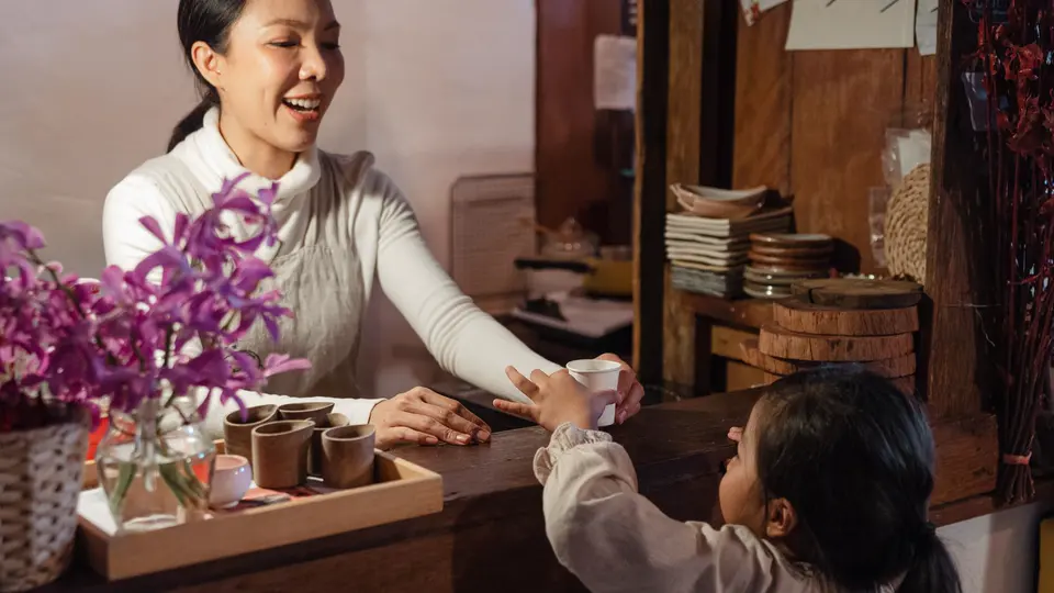 A mother laughs as she hands her young daughter a cup across the counter of a small tea shop, in warm light